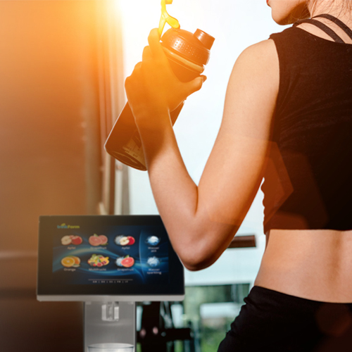 Sportliche Frau füllt ihre Trinkflasche an einem trinkForm® Getränkeautomaten im Fitnessstudio gegen Abendlicht
